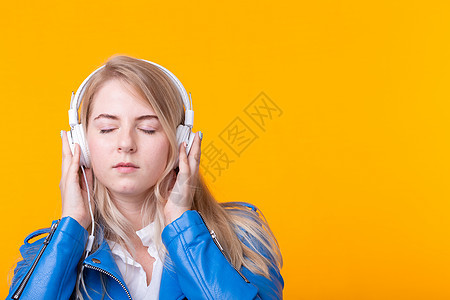 年轻漂亮的金发女学生手持智能手机 头戴蓝色皮夹克耳机 背景为黄色 收听在线广播和音乐订阅的概念知识学生潮人青少年女性商业学校头发图片