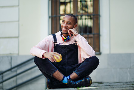 黑人年轻人坐在城市的阶梯上吃苹果背带裤健康衣服快乐牛仔布食物休闲服黑色成人水果图片