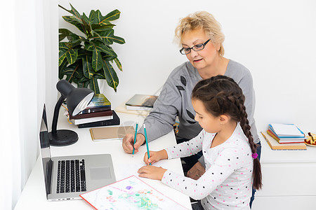 成熟的祖母在家帮助孩子做家庭作业 满意的老奶奶帮助她的孙女在客厅学习 小女孩在笔记本上写字 高级老师坐在她旁边写作教育互联网老太图片