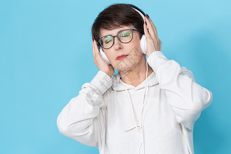美女40 50岁 用大耳机听音乐 蓝底的蓝色背景情绪化女性绿色眼镜女士享受眼皮指甲帽衫棕色图片