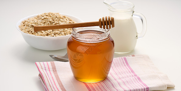 蜂蜜在玻璃透明罐子和白桌上的木棍上 在装有牛奶的挖土机后面图片