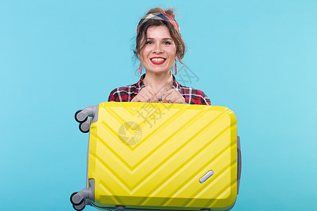 身穿格子衬衫 面带微笑的积极年轻女子拿着一个黄色手提箱 摆在蓝色背景上 旅游和旅行的概念女孩派对流行音乐广告摇滚乐主妇女性口红商图片