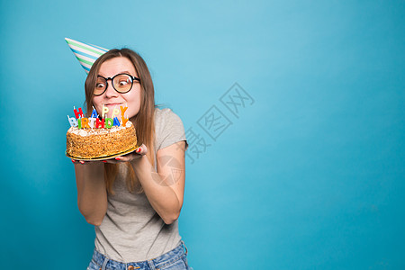 戴着眼镜和贺卡帽的漂亮女孩 手握着一个快乐的生日蛋糕 站在蓝背景上站着 她举起手来工作室蛋糕问候语派对乐趣生日节日甜点艺术学生图片