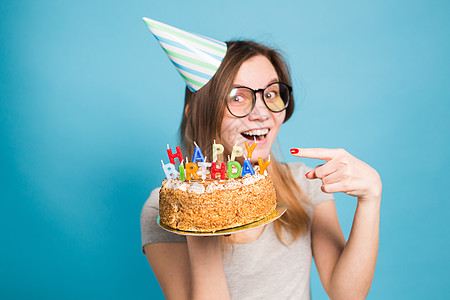 迷人的快乐疯狂的年轻女学生戴着祝贺纸帽 手里拿着一个生日快乐蛋糕 站在蓝色背景下 广告空间风格潮人派对喜悦纪念日节日礼物问候语周图片