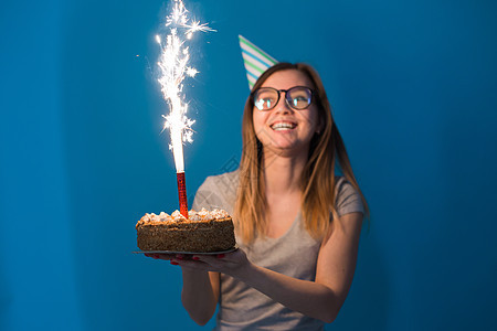 戴着杯子的年轻 模糊的女学生拿着一个祝贺蛋糕和一根蜡烛站在蓝色背景上 生日概念 笑声庆典派对火花情绪周年纪念日艺术娱乐乐趣装饰图片