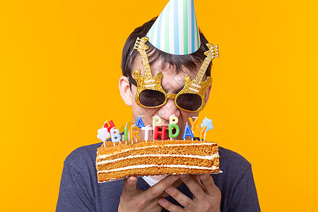 戴着眼镜和纸质祝贺帽子的疯狂快乐的年轻人拿着蛋糕 在黄色背景上站着庆祝生日 欢庆恭喜的概念 掌声 Gibilee庆典惊喜派对娱乐图片