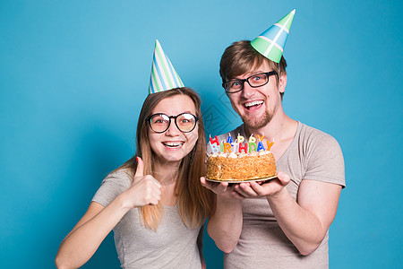 有趣的年轻夫妇戴着纸帽 拿着蛋糕 站在蓝色背景下 做出一张傻脸 祝生日快乐 祝贺和鬼混的概念风格潮人装饰惊喜艺术喜悦节日工作室周图片