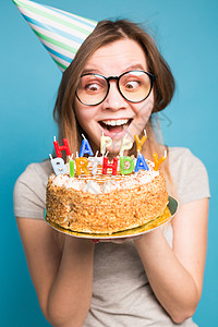 穿着纸帽和眼镜的疯狂可笑女孩 在蓝背景上拿着一个生日蛋糕 恶作剧和问候的概念 笑声女士工作室生日装饰惊喜艺术纪念日庆典风格蓝色图片