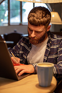 休闲商务人士或自由职业者计划他在笔记本上的工作 在笔记本电脑上用智能手机工作 在咖啡店或家庭办公室的桌子上喝杯咖啡学生男人技术人图片