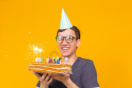 戴着眼镜和纸质祝贺帽子的疯狂快乐的年轻人拿着蛋糕 在黄色背景上站着庆祝生日 欢庆恭喜的概念 掌声 Gibilee派对情绪蓝色娱乐图片