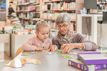外孙女和祖母在市图书馆里拼了个谜题教育架子幸福阅读桌子祖父母学习女孩教学喜悦图片