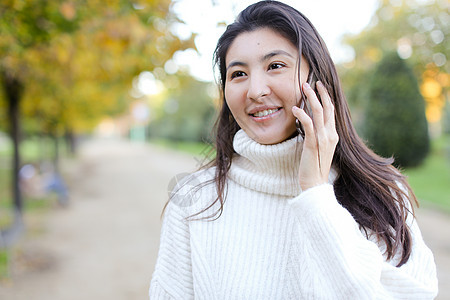 穿着白毛衣的亚洲漂亮女孩 用智能手机说话 在公园散步图片