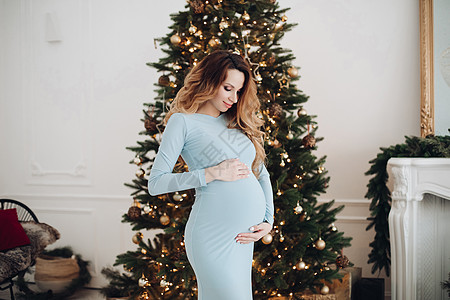 时尚孕妇怀孕的caucasian孕妇在有圣诞树背景的相机上摆姿势背景