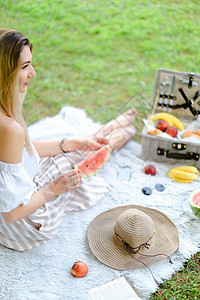 金发女孩坐在水果和帽子旁边 吃西瓜 本底青草的花格上图片