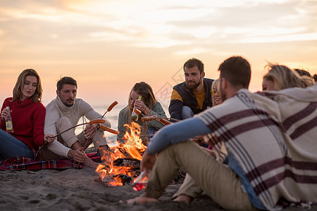 坐在海滩火炉边的年轻朋友团体细胞啤酒饮料青年烹饪温暖篝火香肠友谊野餐背景图片