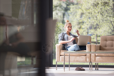 妇女喝咖啡 享受放松的生活方式房间幸福微笑会议屏幕笔记本长椅女性工作互联网背景图片