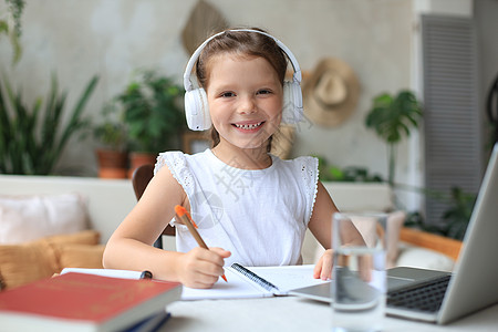 戴着耳机的微笑小女孩在家里使用笔记本电脑在线学习手写 戴着耳机的可爱快乐的小孩在 PC 上上互联网课程或课程书本工作生活方式屏幕图片