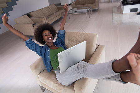 在家里使用笔记本电脑坐在椅子上的非裔美洲妇女技术闲暇活动书柜公寓扶手头发黑发家庭棕色图片