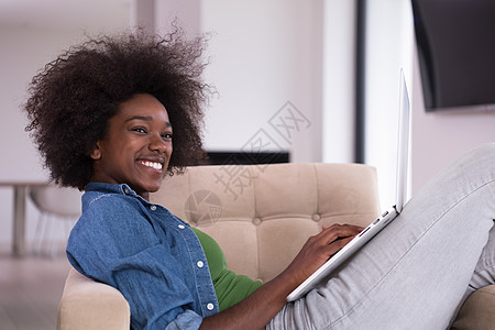在家里使用笔记本电脑坐在椅子上的非裔美洲妇女成人闲暇奢华头发沙发黑发活动书柜互联网学生图片