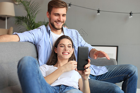 年轻夫妇坐在起居室的沙发上 在智能手机里观看网上内容 笑声男人电话游戏朋友们房间女士家庭乐趣长椅租房者图片