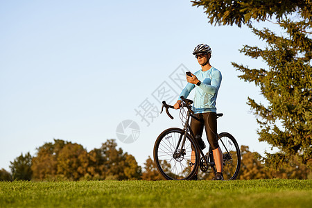 年轻的运动员 穿着运动服和防护头盔的职业自行车手站在公园里骑着自行车 用智能手机检查健身结果图片