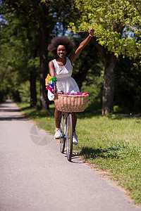 在森林里骑自行车的 年轻漂亮的非洲裔美国女人女性闲暇快乐牛仔裤爆炸晴天女孩衬衫微笑公园图片