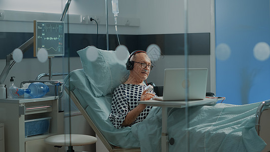 在医院病房使用笔记本电脑和耳机的老年病人图片