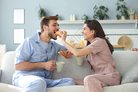 穿着睡衣快乐的一对年轻夫妇在厨房吃早餐 互相喂羊角面包女士鞋垫丈夫妻子食物夫妻长发两个人女性休息图片
