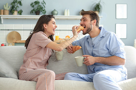 穿着睡衣快乐的一对年轻夫妇在厨房吃早餐 互相喂羊角面包男人黑发谷物长发成年人日常生活杯子鞋垫报纸休息图片
