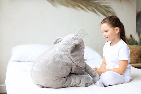 甜美的小女孩坐在她床上 在家中与玩具大象唤醒拥抱微笑房间女性毯子苏醒休息枕头童年图片