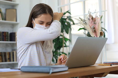 戴医疗保护面罩的女青年在自孤立和隔离期间在家里用笔记本电脑工作 笑声流感症状工人感染药品女士办公室疾病笔记商业图片