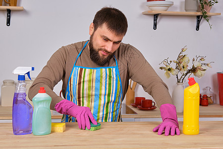 厨房里长着胡子的年轻英俊男子 试图用洗涤剂 刷子 喷雾器打扫桌子手套橡皮围裙蓝色看门人画笔丈夫男性家务家政图片