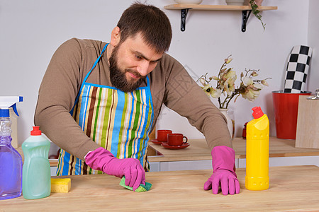 厨房里长着胡子的年轻英俊男子 试图用洗涤剂 刷子 喷雾器打扫桌子蓝色家庭橡皮家务工作清洁工画笔抹布服务用品图片