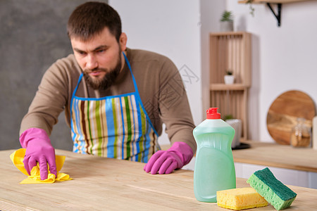 厨房里长着胡子的年轻英俊男子 试图用洗涤剂 刷子 喷雾器打扫桌子家政看门人家庭橡皮围裙蓝色服务卫生丈夫喷雾图片