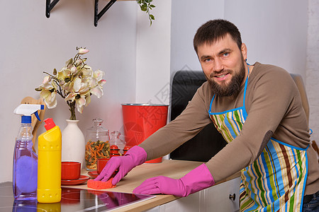 厨房的英俊长胡子男 身着围裙和粉色手套 用洗涤剂清洗烹饪表面男性快乐公寓卫生用品桌子火炉胡须灶台家政图片