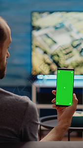 白种人垂直持有绿色屏幕智能手机 并用绿屏互联网电视房间长椅男人桌面闲暇工作技术商业图片