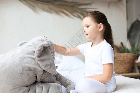 甜美的小女孩坐在她床上 在家中与玩具大象微笑拥抱枕头童年卧室毯子女孩男人女性唤醒图片