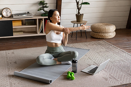 年青的亚洲健身女孩做伸展 在家运动锻炼 伸臂和看笔记本电脑 在计算机上跟踪练习录像运动员活力女士瑜伽运动装房子冥想哑铃有氧运动身图片