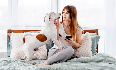 狗睡觉床上带着狗和智能手机的女孩卧室休息微笑电话动物说谎朋友宠物乐趣幸福背景