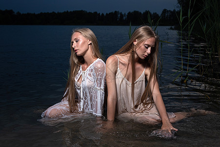 夏天晚上 两个年轻的双姐妹在湖水中穿着轻礼服姐姐裙子天空青少年冒充金发海滩手电筒池塘双胞胎图片