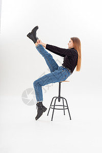 身穿黑色高领 蓝色牛仔裤的年轻有吸引力的caucasia女人穿上靴子凳子冒充姿势成人长发金发椅子天幕头发工作室图片