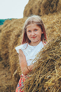 一个穿着粉红裙子的金发女学生 在炎热的夏日 爬上一大堆稻草褐色天空冒充太阳小麦闲暇蓝色青年草垛女孩图片