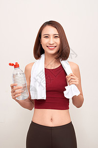 带着一瓶水和毛巾的运动女孩 肩上戴着轻光背景的便衣瓶子身体女性白色健身房讲师图片