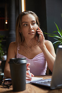 坐在咖啡馆用笔记本电脑坐着的年轻美丽的年轻女士 由智能手机打电话学生电话桌子企业家工具成人晚餐咖啡自由职业者技术图片
