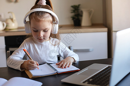 戴耳机的小女孩坐在办公桌前在笔记本上写字在线学习在家做运动 小孩子手写准备隔离作业 上网络课或在室内上课生活方式书本网站小书房知图片