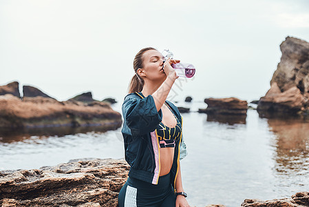 水就是生命 穿着运动服的运动型年轻女子坐在沙滩上的石头上喝水图片