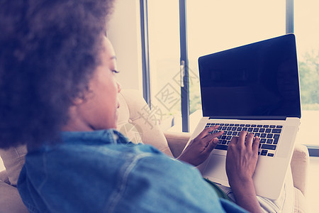 在家里使用笔记本电脑坐在椅子上的非裔美洲妇女监视器技术学生成人黑发书柜棕色长椅活动客厅图片