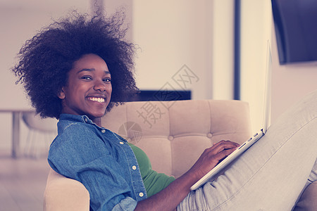 在家里使用笔记本电脑坐在椅子上的非裔美洲妇女学生棕色化妆品公寓监视器沙发客厅技术闲暇女性图片