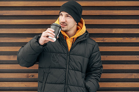 身着冬季衣服的青年男子站在咖啡边 在街上与木墙对撞幸福男性快乐喜悦杯子青少年男人微笑女孩饮料图片