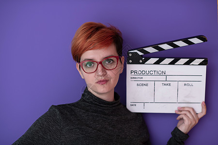 红发女人拿着紫色背景的电影拍扣相机生产商业乐趣视频制片人微笑摄影黑板行动图片